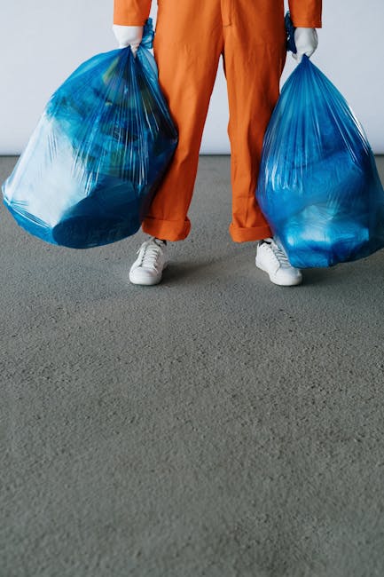 A person standing indoors on a grey carpeted floor is holding two large blue plastic rubbish bags, one in each hand. The individual is dressed in orange overalls and white sneakers. The bags are fille